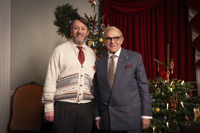 David Mitchell and Sir David Suchet stand indoors in front of two decorated Christmas trees; the person on the left wears a patterned cardigan with a red tie, and the person on the right wears a grey pinstripe suit. The festive setting includes ornaments, candles, red curtains and warm Christmas décor.
