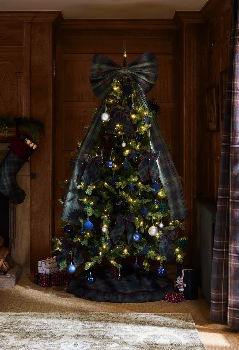 A Christmas tree decorated with blue and silver baubles, warm white lights and large plaid bows stands in a cosy wood‑panelled room. Wrapped presents sit beneath the tree, with a matching plaid stocking hanging from the nearby fireplace and coordinating plaid curtains completing the festive scene.