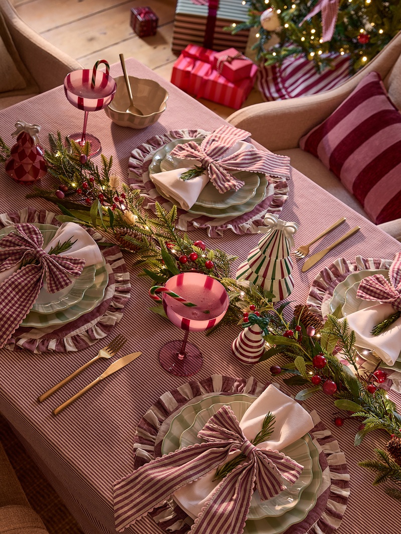 A Christmas dining table decorated with red, pink and green festive place settings, striped bows, scalloped plates and pink goblets. An evergreen garland with berries and cone‑shaped decorations runs along the centre, with a lit Christmas tree and wrapped presents in the background.