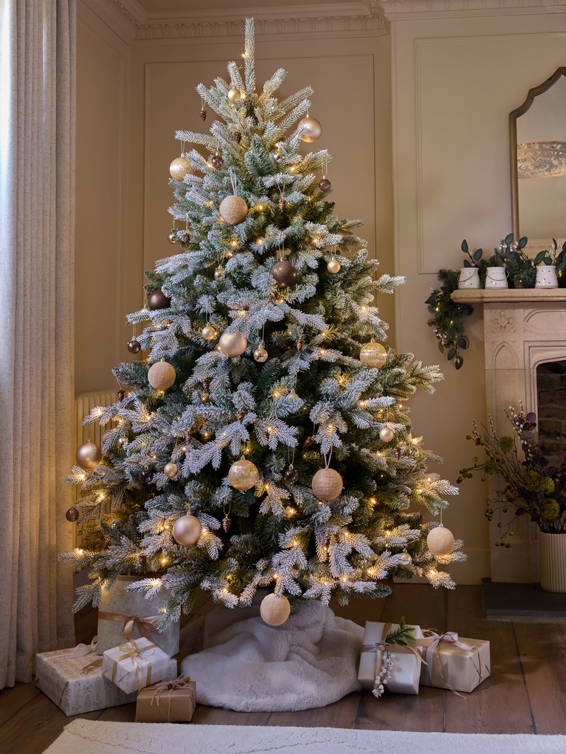 A frosted Christmas tree decorated with warm white lights and gold, bronze and champagne ornaments stands beside a festive fireplace. Wrapped neutral‑toned gifts sit on a soft white tree skirt, with garland, candles and a mirror on the mantel completing the elegant holiday scene.
